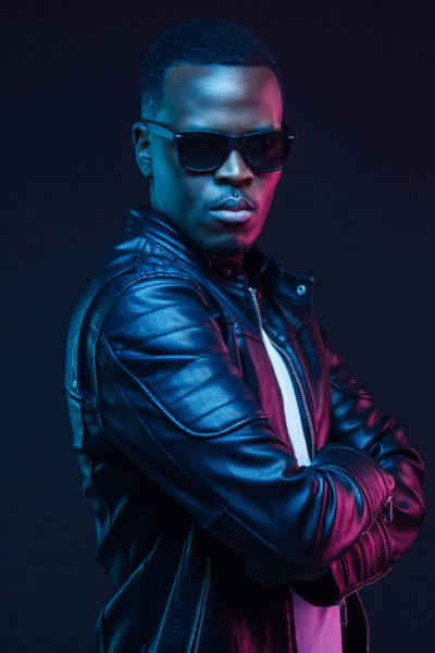 Studio neon portrait of stylish black man standing with crossed arms, wearing leather jacket and sunglasses Studio neon portrait of stylish black man standing with crossed arms, wearing leather jacket and sunglasses