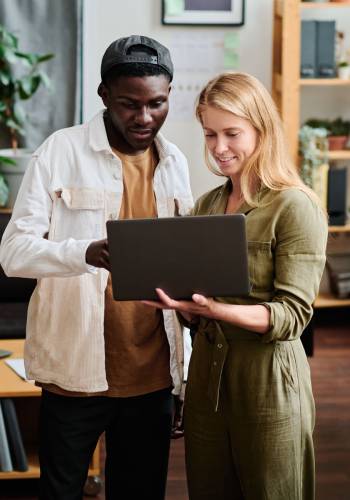 Two young intercultural employees in casualwear looking at screen of laptop Two young intercultural employees in casualwear looking at screen of laptop held by blond businesswoman during discussion