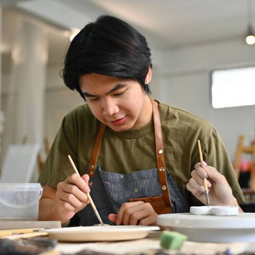 Two teenage Asian students focused on painting ceramic pieces in an art classroom together Two teenage Asian students focused on painting ceramic pieces in an art classroom together.